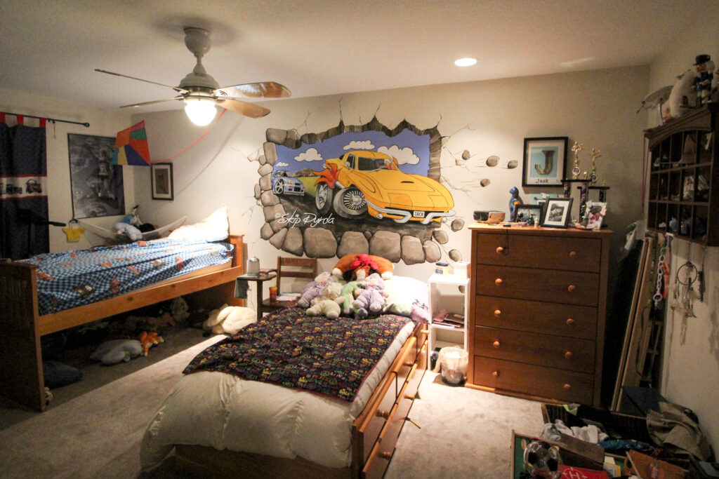 Photo of a wall mural of a cartoon style image of a big yellow corvette being chased by a BMW, by Skip Dyrda. Full room view